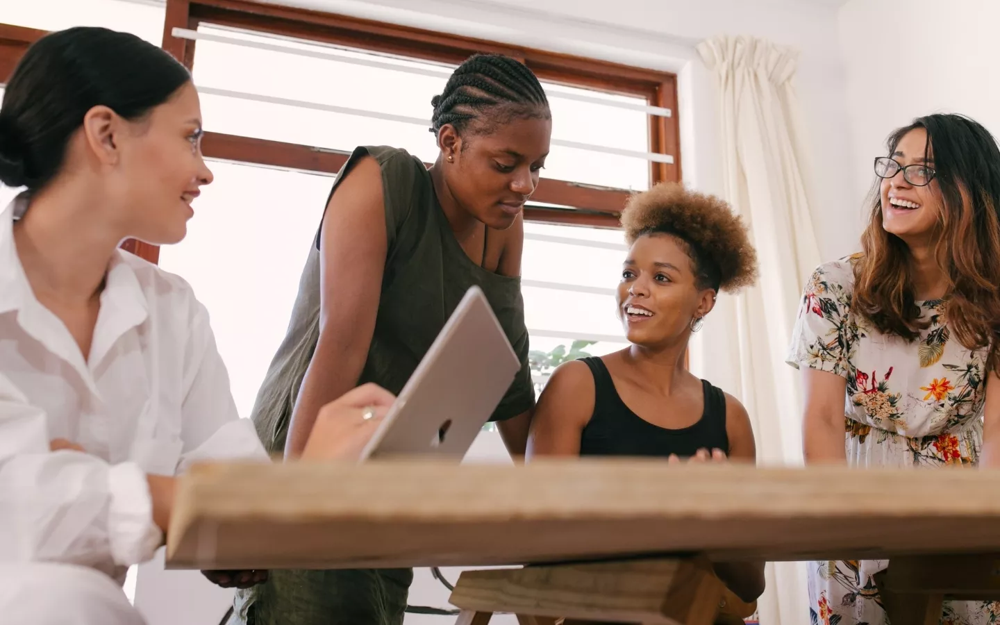 Four women engaged in a conversation and collaboration in a bright, casual workspace with a laptop on the table.
