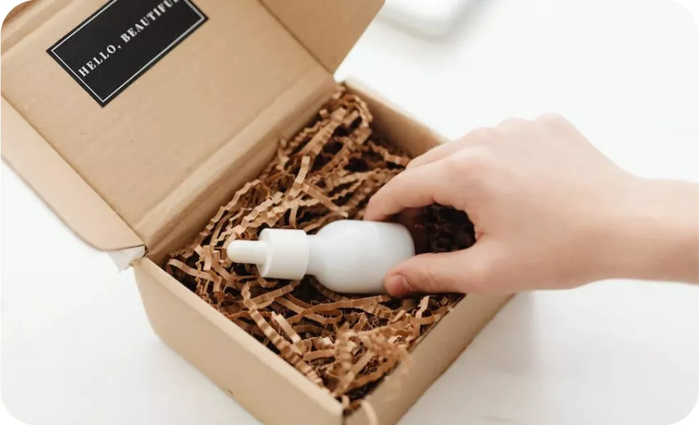 Hand pulling a white dropper bottle from a cardboard box filled with packing paper.