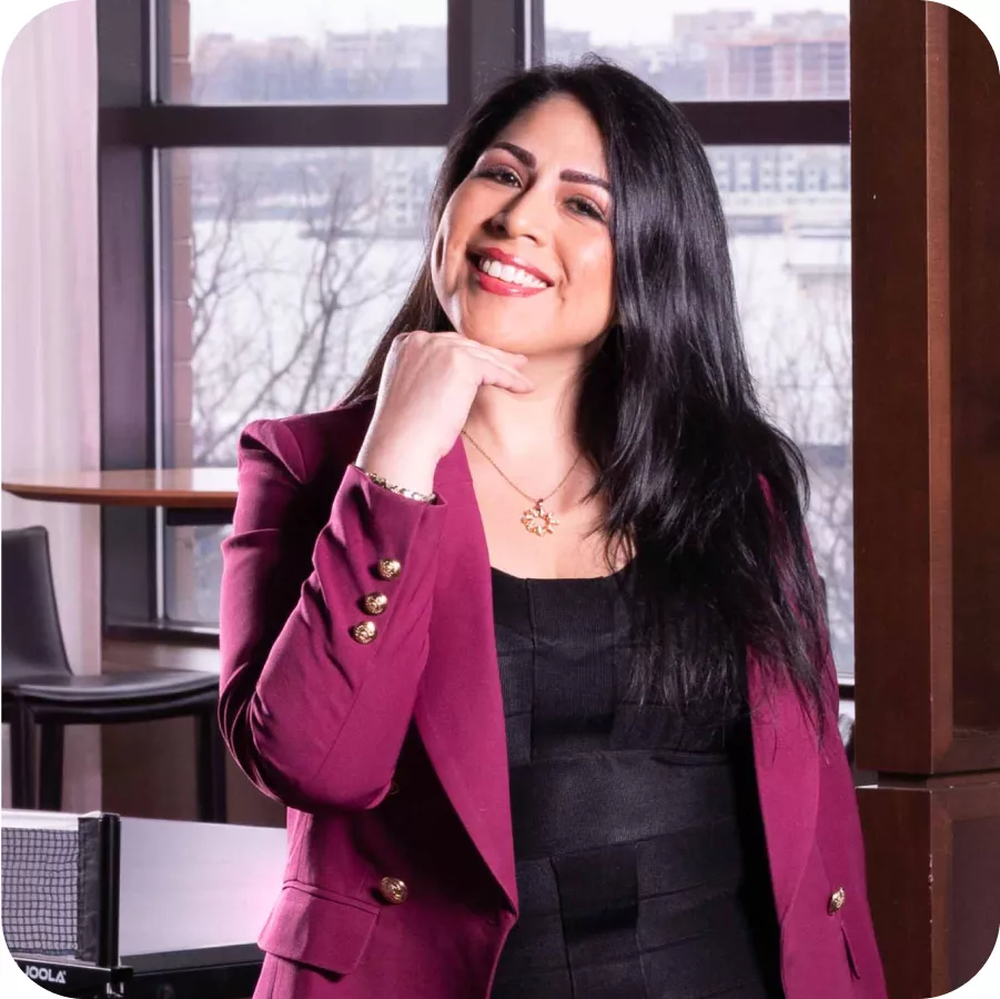 Magaly Ponce smiling and posing with a hand on her chin, wearing a burgundy blazer and standing in front of a window.