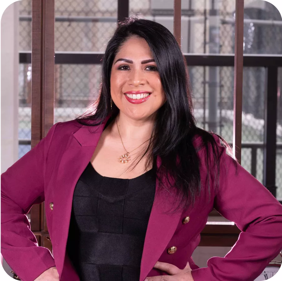 Magaly Ponce smiling confidently in a burgundy blazer, standing with hands on hips in front of a window.